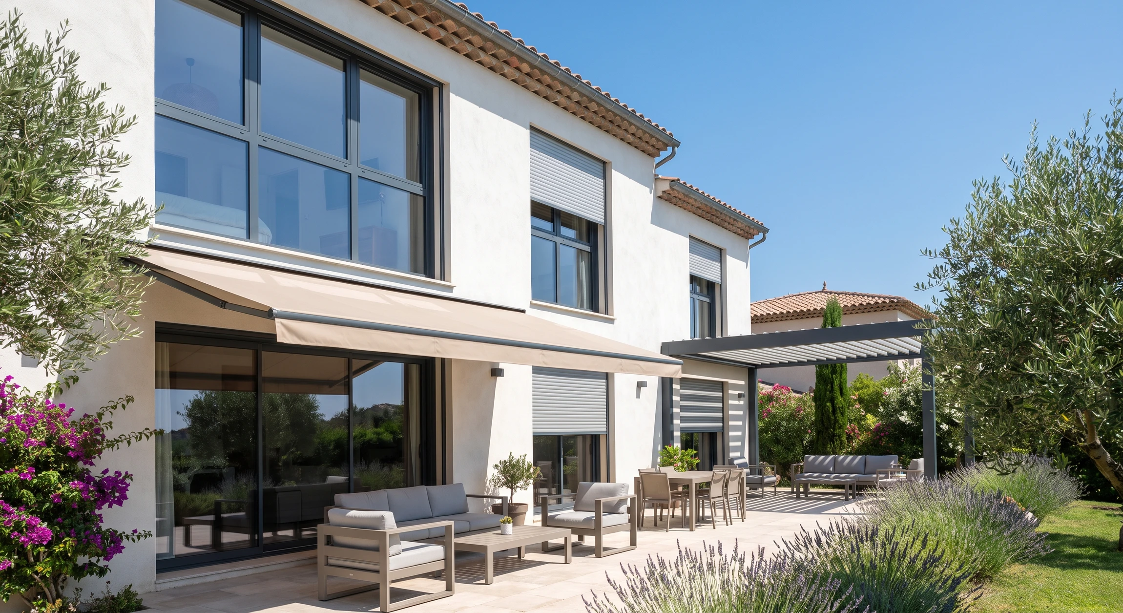 Photographie d'une façade résidentielle méditerranéenne équipée de multiples solutions d'installation : fenêtres PVC grand format avec menuiseries aluminium gris anthracite, store banne motorisé déployé sur toile beige, volets roulants en aluminium semi-abaissés, pergola bioclimatique à lames orientables en arrière-plan. Terrasse aménagée visible avec salon de jardin. Environnement ensoleillé, végétation paysagère, mettant en avant la qualité et la diversité des interventions de l'entreprise dans le sud de la France.