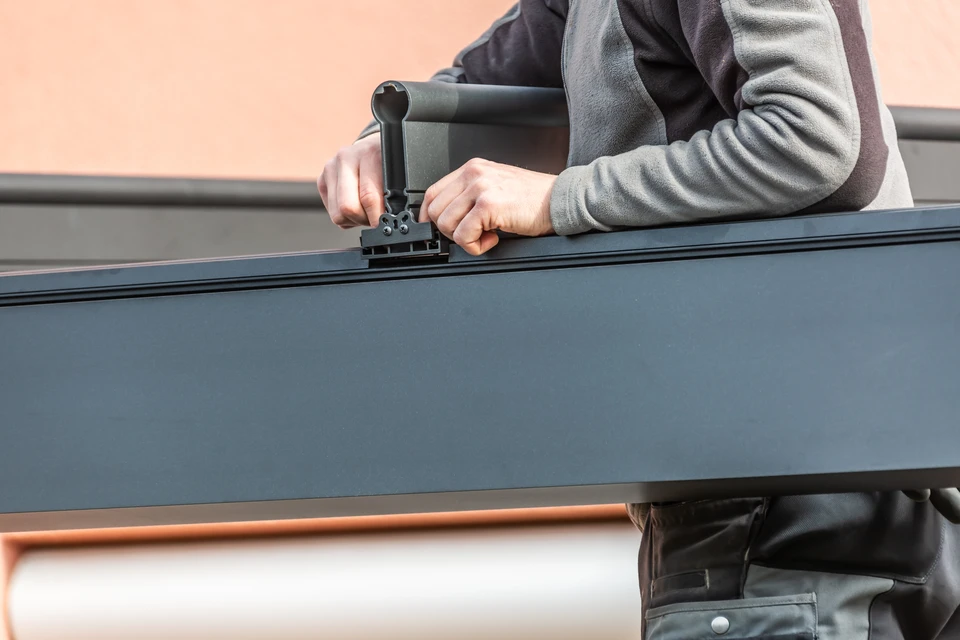 Terrasse extérieure résidentielle moderne équipée d'une pergola bioclimatique en aluminium anthracite avec lames orientables horizontales, partiellement ouvertes pour créer une zone d'ombre. La structure est adossée à une maison contemporaine. Un mobilier de jardin minimal est visible (chaises, table basse). Le sol est en dalles de béton clair. Le ciel est dégagé. La photo met en avant la qualité de l'installation, la précision des finitions, et l'intégration harmonieuse de la pergola dans l'environnement résidentiel.