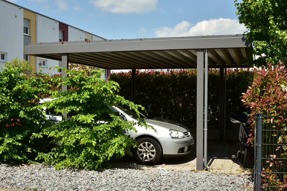 Carport moderne en structure aluminium anthracite avec toit plat, abritant un véhicule personnel. La structure repose sur des poteaux carrés, avec un sol en pavés de béton. La couverture protège le véhicule de la pluie, du soleil et des UV. Environnement résidentiel avec façade de maison et végétation en arrière-plan. Perspective frontale montrant la protection offerte et l'élégance de la structure.