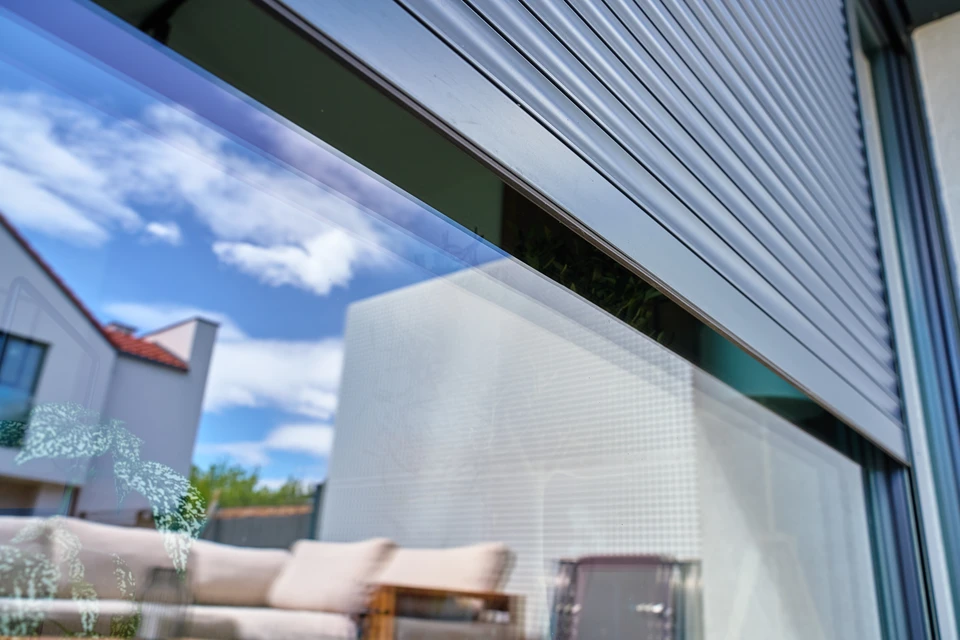 Façade extérieure d'une maison résidentielle moderne avec une fenêtre ou baie vitrée équipée d'un volet roulant motorisé en aluminium anthracite, partiellement ou totalement descendu, montrant clairement le coffre de volet roulant en tête de fenêtre. Menuiserie aluminium gris moderne. Installation en applique sur enduit blanc ou clair. Environnement résidentiel calme avec jardin visible. Photo réaliste en lumière naturelle de jour.
