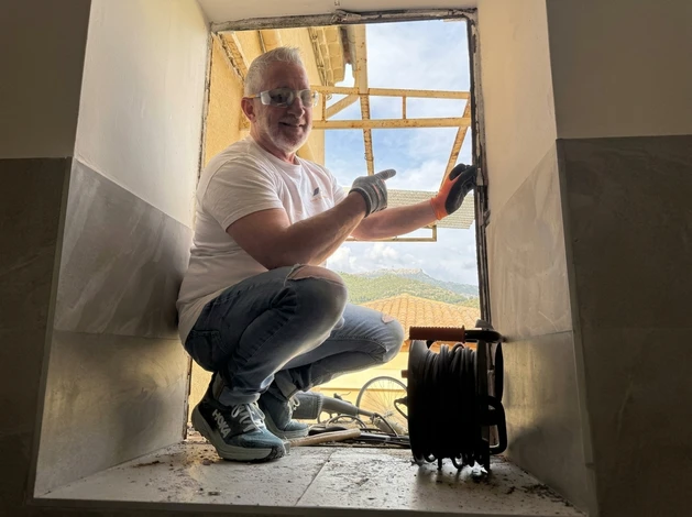 Photographie réelle en extérieur d'un artisan menuisier/installateur (Serge Grasso ou un technicien confirmé de l'entreprise) travaillant à l'installation d'une fenêtre PVC ou d'un élément de fermeture extérieure sur une façade résidentielle. L'artisan est concentré, professionnel, portant équipement de sécurité approprié (gants, lunettes si pertinent). En arrière-plan, on voit la maison du client, un environnement méditerranéen résidentiel, ciel dégagé. La composition doit transmettre la précision, la confiance et l'expertise. Éviter tout cliché générique, favoriser une ambiance authentique de travail de qualité réalisé sur le terrain.